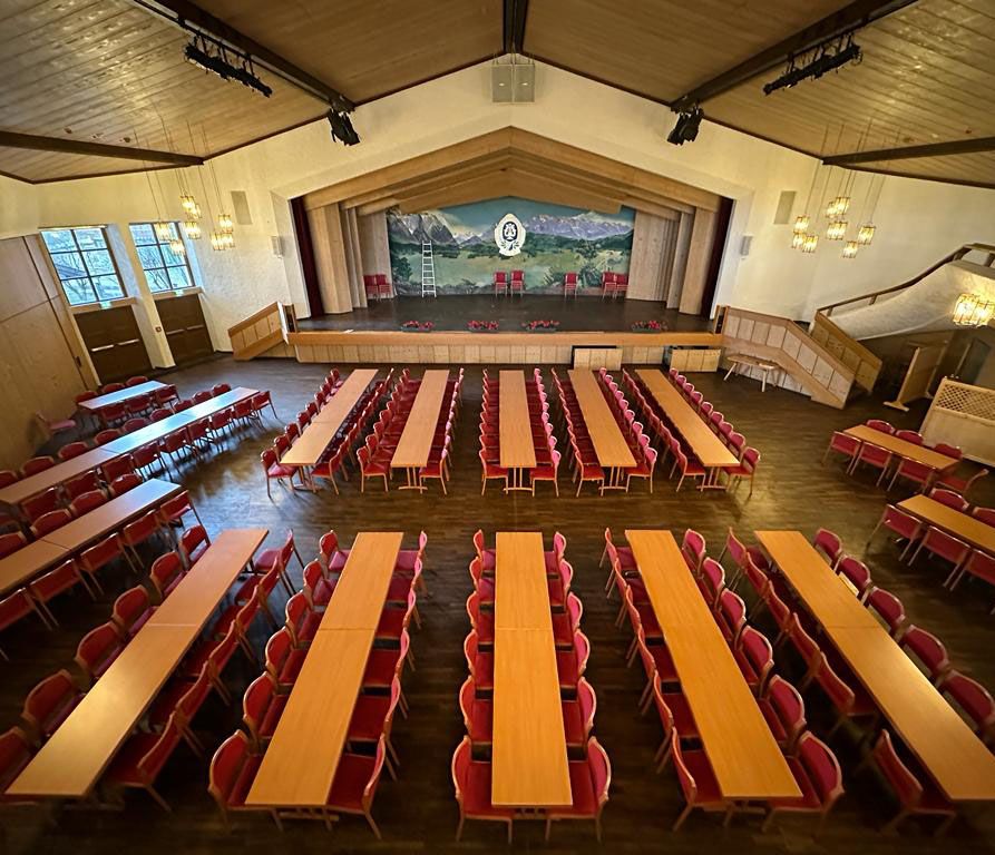 Großer Saal mit Bühne, mehreren langen Tischen und roten Stühlen, Holzdecke und Kronleuchtern