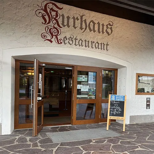 Eingang zum Kurhaus Restaurant mit offenem Holztürrahmen, zwei beschrifteten Tafeln und Laterne an weißer Wand