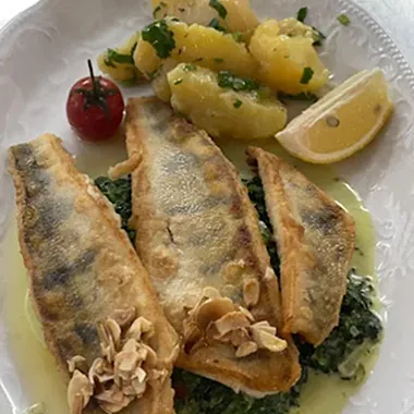 Drei gebratene Fischfilets auf einem Bett aus grünem Blattgemüse, garniert mit Mandelblättchen. Daneben liegen gekochte Kartoffeln, eine halbe Zitrone und eine Kirschtomate.
