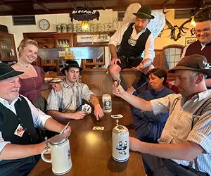 Eine Gruppe von sechs Personen in traditioneller Kleidung sitzt um einen Tisch in einer Gaststätte. Eine Person steht auf dem Tisch und hält ein Bein auf der Tischkante. Auf dem Tisch stehen Bierkrüge.