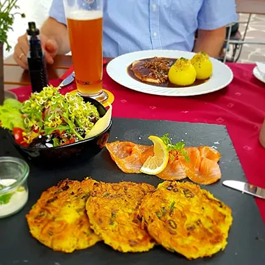 Drei goldbraune Kartoffelpuffer auf einem schwarzen Teller, daneben Lachs mit Zitronenscheibe und ein Salat in einer schwarzen Schüssel. Im Hintergrund ein Glas Bier und ein Teller mit zwei Kartoffelknödeln und einer dunklen Soße.