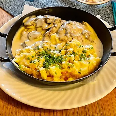 Schwarze Pfanne mit zwei Portionen: oben cremige Pilze, unten knusprige Käsespätzle, garniert mit frischem Schnittlauch.