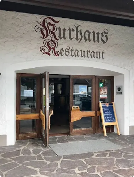 Eingang zum Kurhaus Restaurant mit offenen Holztüren, einem Schild mit der Aufschrift 'Willkommen' und Steinboden.