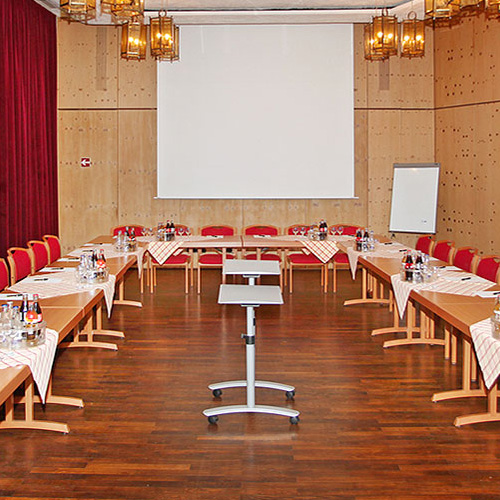 Seminarraum mit U-förmiger Anordnung von Holztischen, roten Stühlen, Tischdecken, Getränkeflaschen, einem weißen Projektionsschirm und einem Flipchart.