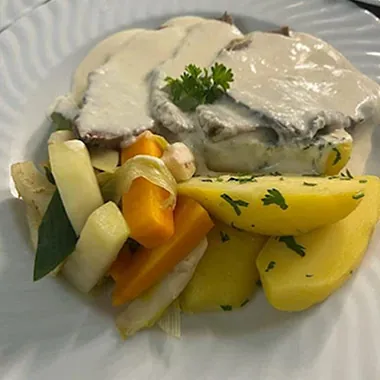 Ein Teller mit zwei Scheiben Fleisch in einer cremigen Sauce, garniert mit einem Blatt Petersilie. Dazu liegen gekochte Kartoffeln und eine bunte Mischung aus Gemüse, darunter Karotten, Zucchini und Lauch.