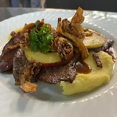 Ein Teller mit püriertem Kartoffelbrei, Scheiben von Fleisch, Äpfeln und knusprigen Zwiebelringen, garniert mit frischem Koriander.