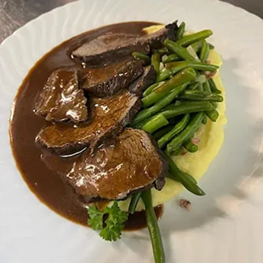Scheiben von Rinderbraten auf einem Bett aus Kartoffelpüree, garniert mit grünen Bohnen und einer dunklen Soße. Ein Blatt Koriander ist sichtbar.