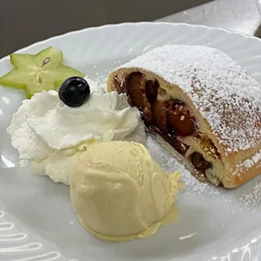Ein Stück Apfelstrudel mit Puderzucker, einer Kugel Vanilleeis, Sahne und einer Sternfrucht, garniert mit einer Heidelbeere.