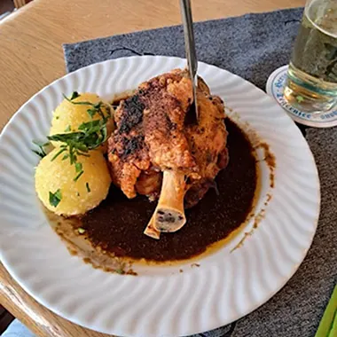 Ein Teller mit einem gebratenen Haxenstück, garniert mit einer Gabel, umgeben von zwei Kartoffelknödeln und einer dunklen Soße. Im Hintergrund steht ein Glas Bier.