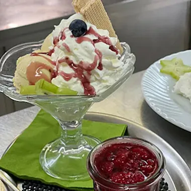 Eisbecher mit Sahne, einer Waffel, Blaubeeren und Melonenstücken, serviert in einem Glas auf einem grünen Tuch. Daneben ein Glas mit roter Fruchtsoße.