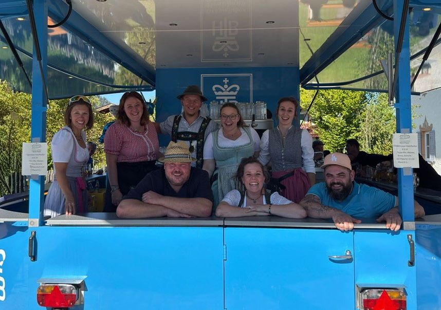 Mehrere Personen in traditioneller Tracht stehen und sitzen in einem offenen blauen Fahrzeug mit HB-Logo, umgeben von Bäumen und Gebäuden.
